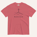 A red short-sleeve crewneck t-shirt featuring a black line art graphic of a cross on a hill with the word 'Redeemed' printed underneath, displayed on a light cream background.
