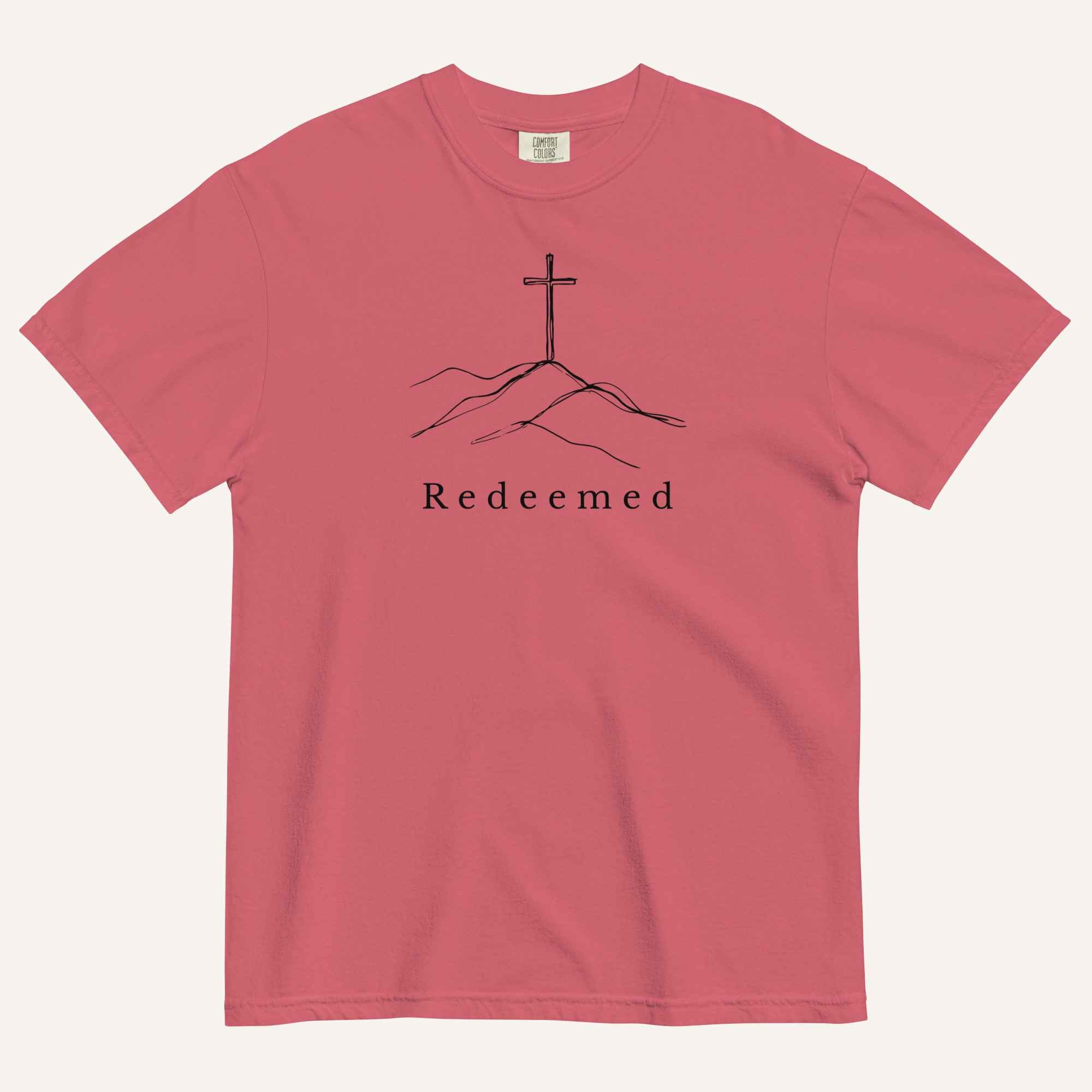 A red short-sleeve crewneck t-shirt featuring a black line art graphic of a cross on a hill with the word 'Redeemed' printed underneath, displayed on a light cream background.
