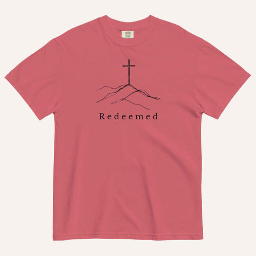 A red short-sleeve crewneck t-shirt featuring a black line art graphic of a cross on a hill with the word 'Redeemed' printed underneath, displayed on a light cream background.