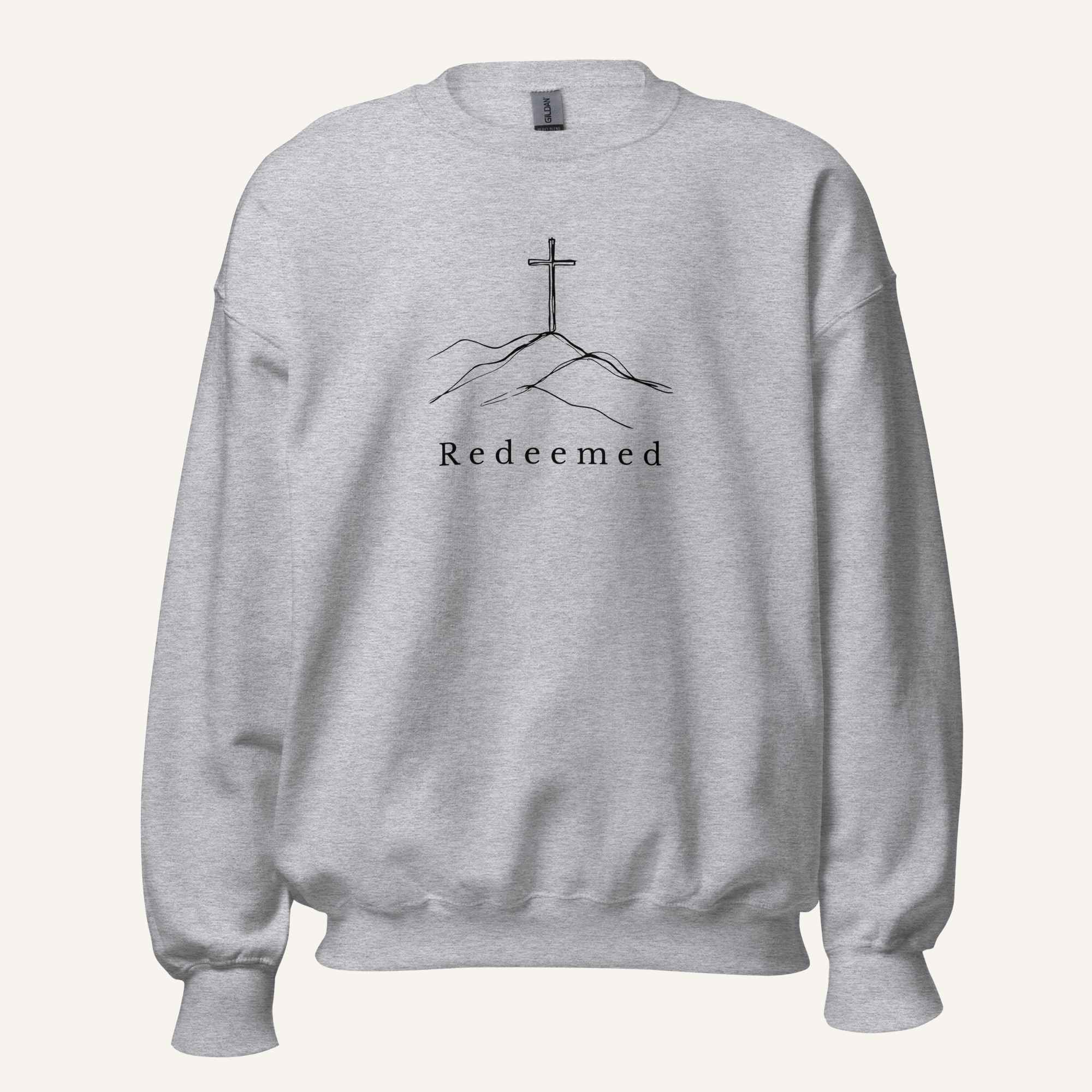 A light heather gray crewneck sweatshirt featuring a black graphic of a cross on a hill with the word 'Redeemed' printed below it, displayed on a clean off-white background.