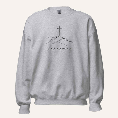 A light heather gray crewneck sweatshirt featuring a black graphic of a cross on a hill with the word 'Redeemed' printed below it, displayed on a clean off-white background.