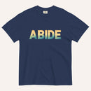 A navy blue short-sleeve t-shirt with the word 'ABIDE' printed across the chest in a bold sans-serif font. The letters feature a vertical gradient transitioning from yellow to light blue and teal.
