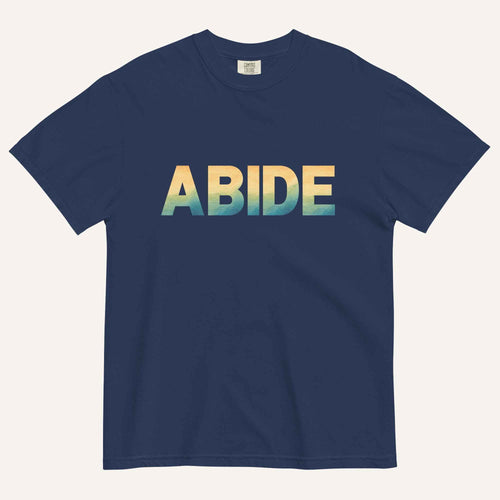 A navy blue short-sleeve t-shirt with the word 'ABIDE' printed across the chest in a bold sans-serif font. The letters feature a vertical gradient transitioning from yellow to light blue and teal.
