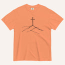 A peach-colored short-sleeve crewneck t-shirt featuring a minimalist black line art graphic of a cross on a hill, displayed flat against a light off-white background.