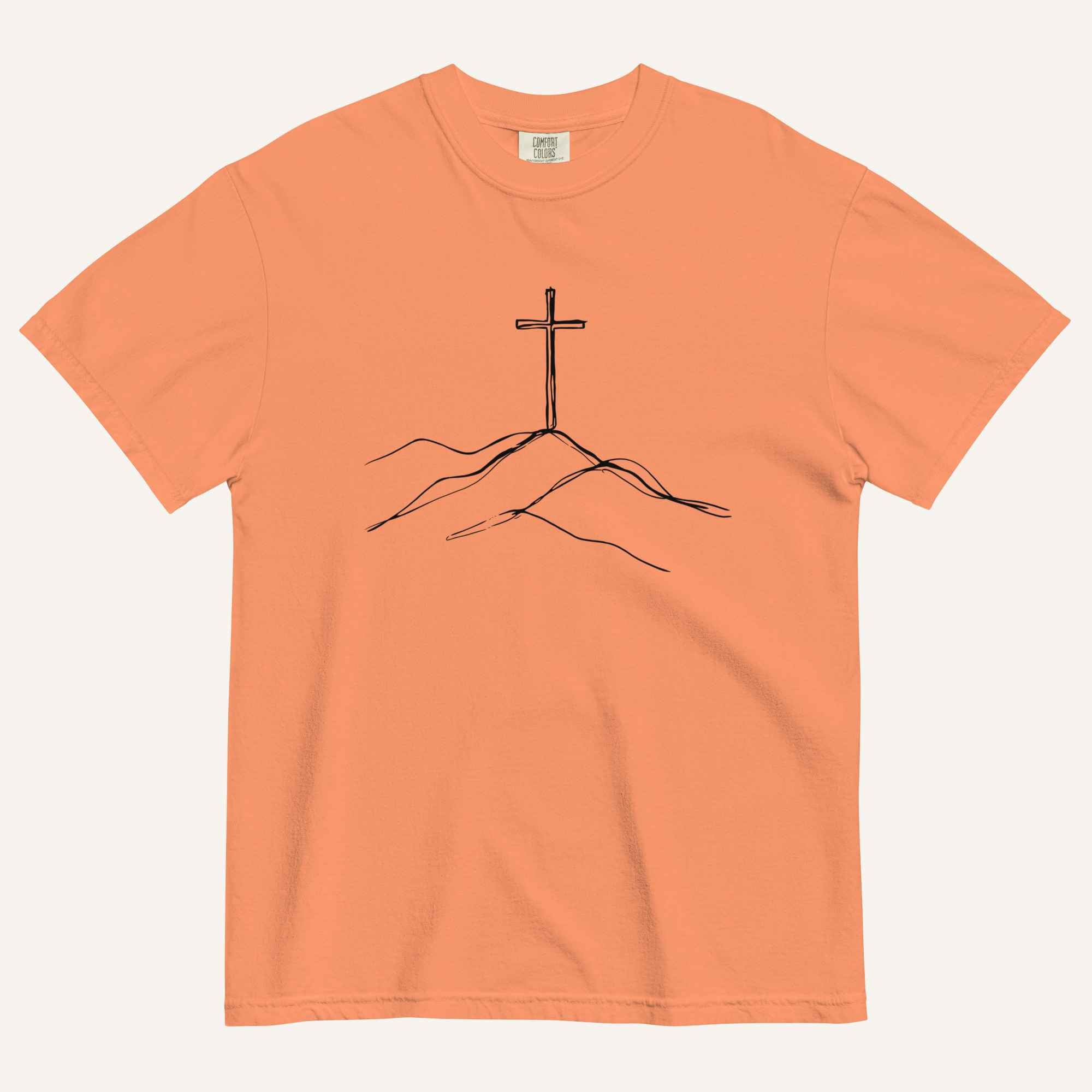 A peach-colored short-sleeve crewneck t-shirt featuring a minimalist black line art graphic of a cross on a hill, displayed flat against a light off-white background.