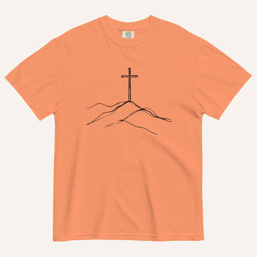 A peach-colored short-sleeve crewneck t-shirt featuring a minimalist black line art graphic of a cross on a hill, displayed flat against a light off-white background.