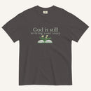 Dark gray t-shirt with 'God is still writing your story' text and book graphic on a white background