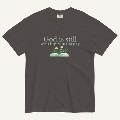Dark gray t-shirt with 'God is still writing your story' text and book graphic on a white background