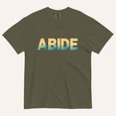 An olive green short-sleeve t-shirt with the word 'ABIDE' printed across the chest in a horizontal gradient transitioning from yellow to light blue and teal, displayed on a light cream background.