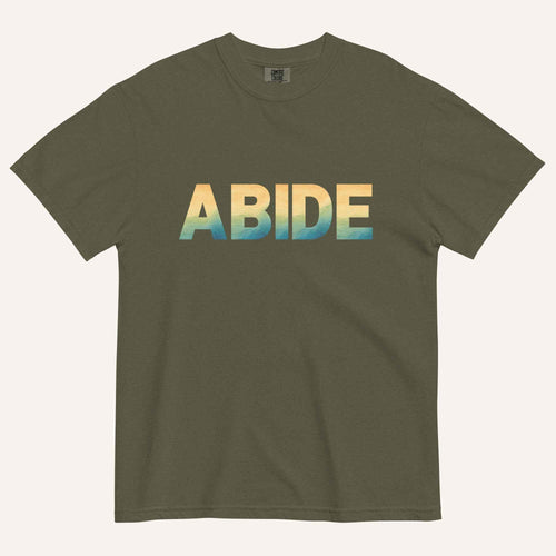 An olive green short-sleeve t-shirt with the word 'ABIDE' printed across the chest in a horizontal gradient transitioning from yellow to light blue and teal, displayed on a light cream background.