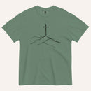 A green short-sleeve crewneck t-shirt featuring a minimalist black line art graphic of a cross rising from hills on the chest, displayed flat against a light cream background.