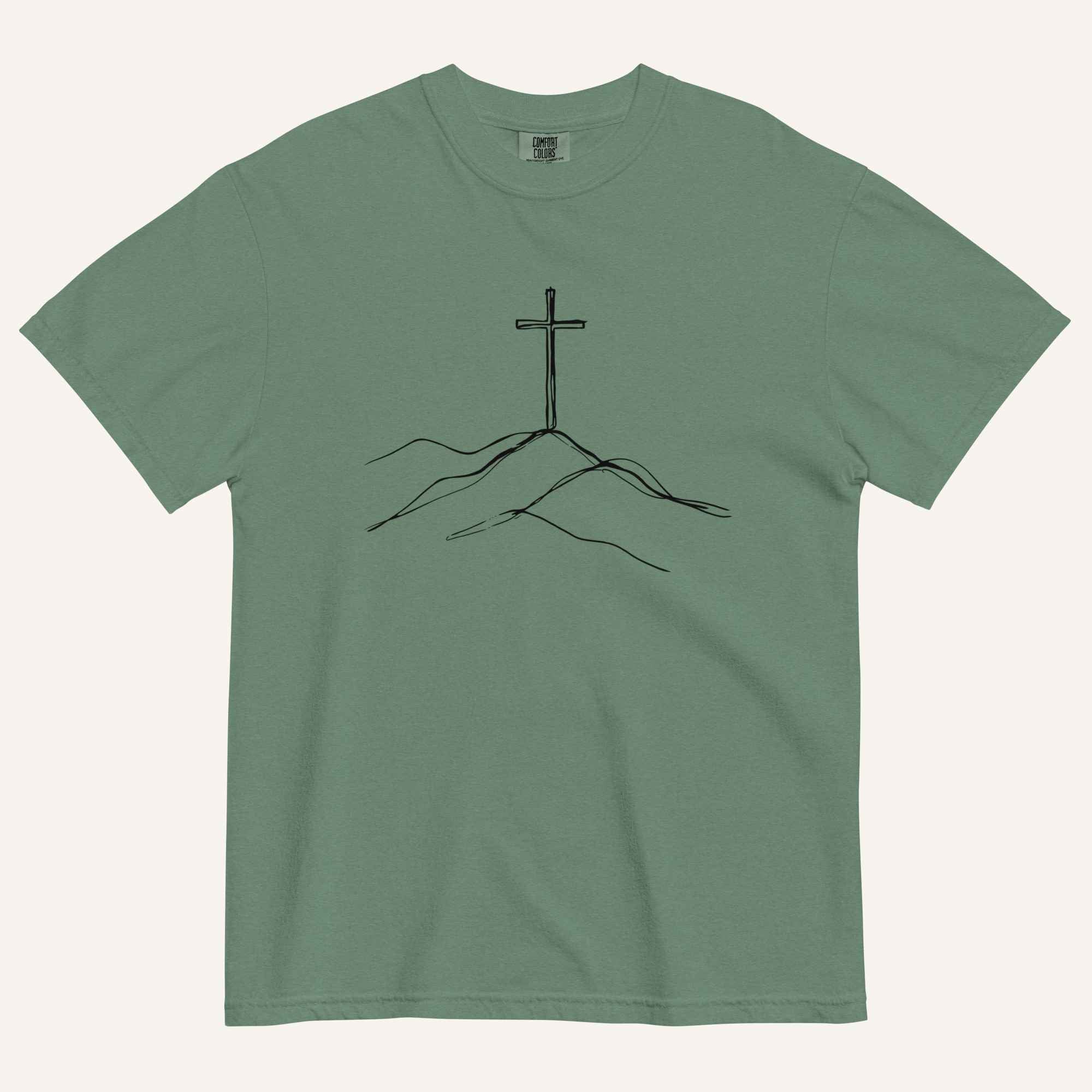 A green short-sleeve crewneck t-shirt featuring a minimalist black line art graphic of a cross rising from hills on the chest, displayed flat against a light cream background.