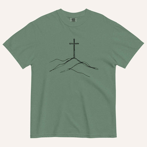 A green short-sleeve crewneck t-shirt featuring a minimalist black line art graphic of a cross rising from hills on the chest, displayed flat against a light cream background.