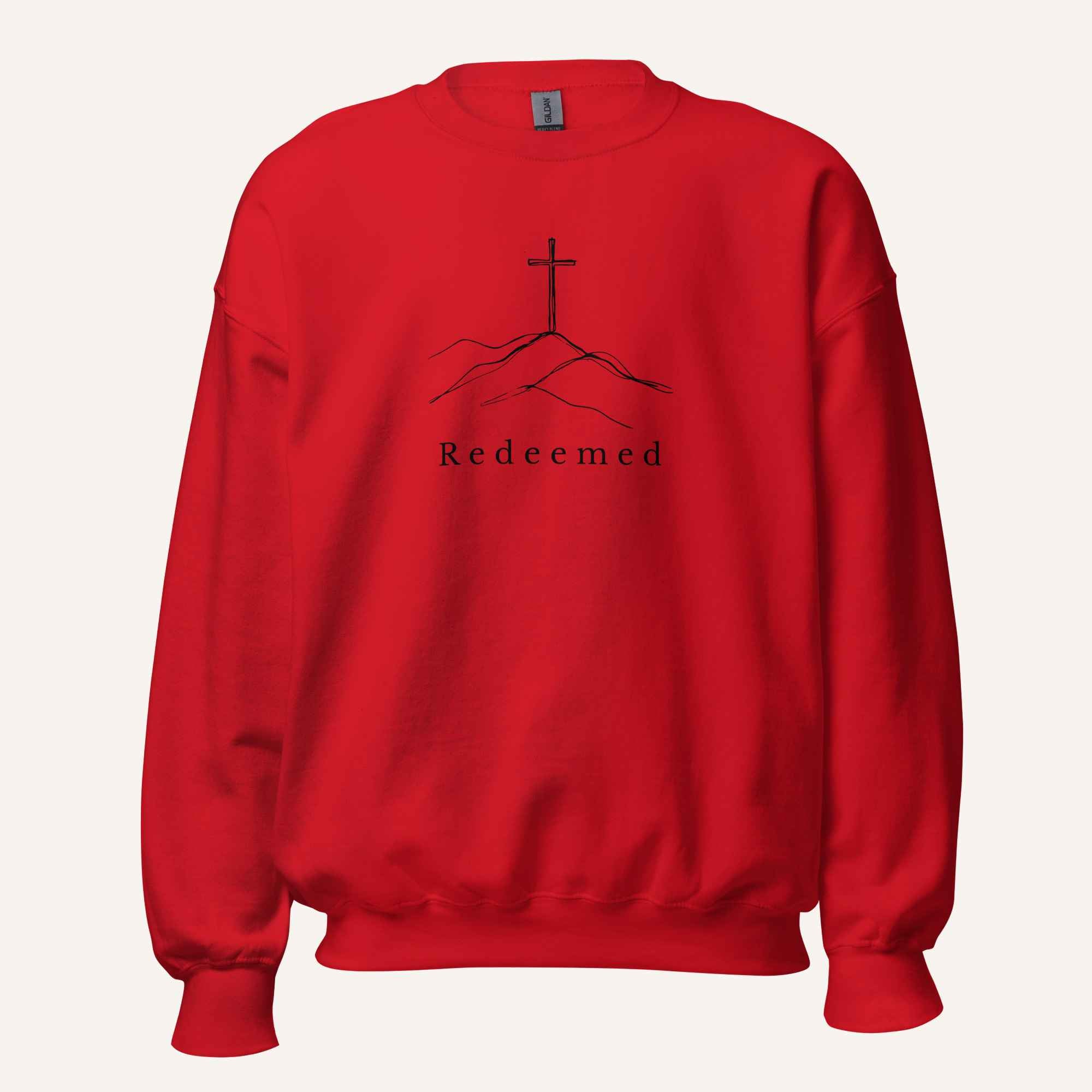 A bright red crewneck sweatshirt featuring a black graphic of a cross on a hill with the word 'Redeemed' printed below it, displayed on a clean off-white background.