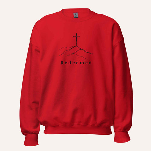 A bright red crewneck sweatshirt featuring a black graphic of a cross on a hill with the word 'Redeemed' printed below it, displayed on a clean off-white background.