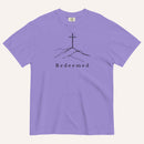 A purple short-sleeve crewneck t-shirt featuring a black line art graphic of a cross on a hill with the word 'Redeemed' printed underneath, displayed on a light cream background.