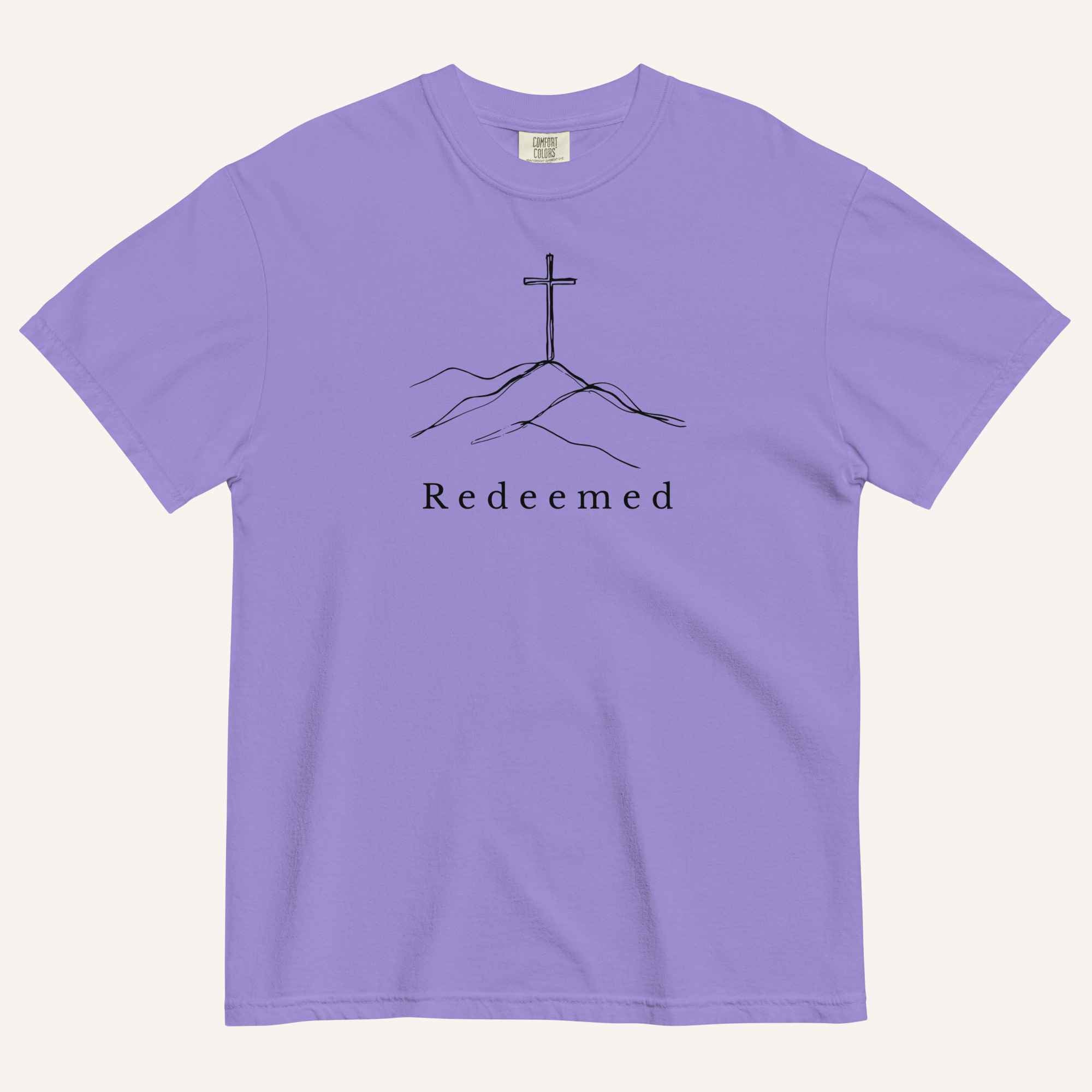 A purple short-sleeve crewneck t-shirt featuring a black line art graphic of a cross on a hill with the word 'Redeemed' printed underneath, displayed on a light cream background.