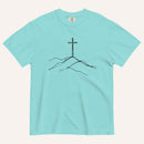 A light aqua blue short-sleeve crewneck t-shirt with a minimalist black line art graphic of a cross on a hill, displayed flat against a light cream background.