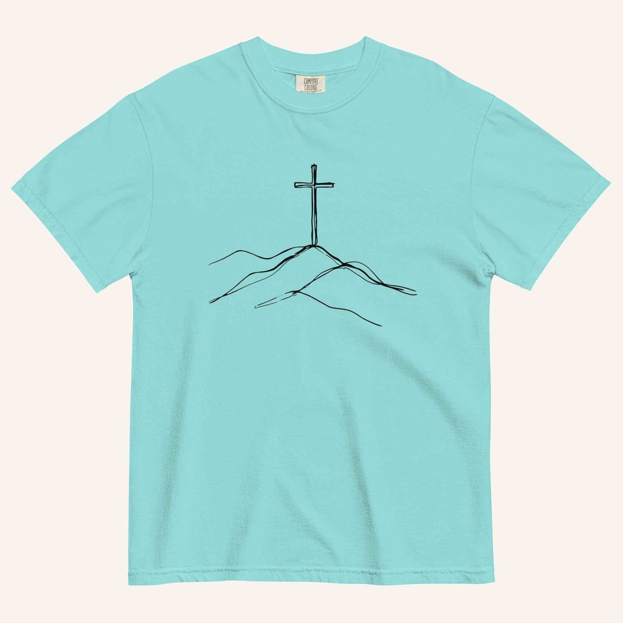 A light aqua blue short-sleeve crewneck t-shirt with a minimalist black line art graphic of a cross on a hill, displayed flat against a light cream background.