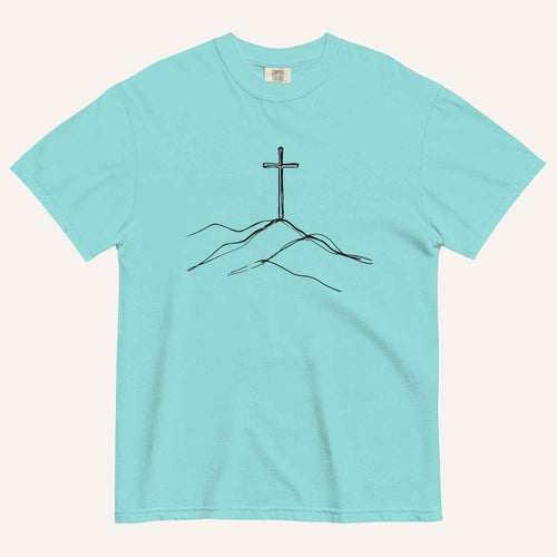 A light aqua blue short-sleeve crewneck t-shirt with a minimalist black line art graphic of a cross on a hill, displayed flat against a light cream background.