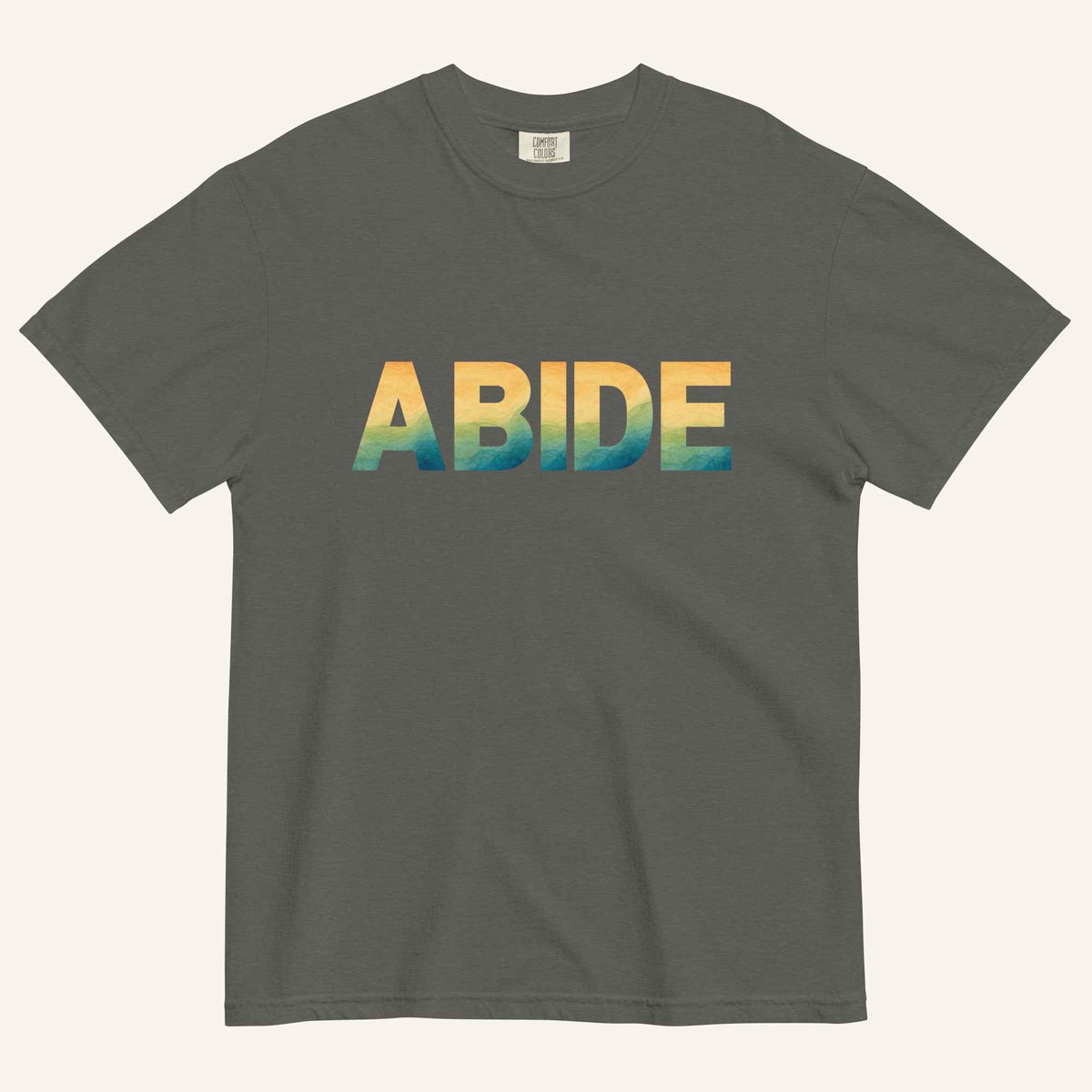 A dark gray crewneck t-shirt featuring the word 'ABIDE' printed across the chest in a textured, multi-colored gradient pattern of yellow, orange, and blue, displayed on a light cream background.