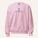 A light pink crewneck sweatshirt featuring a black line art graphic of a cross on top of simple mountains on the chest, displayed against a solid off-white background.
