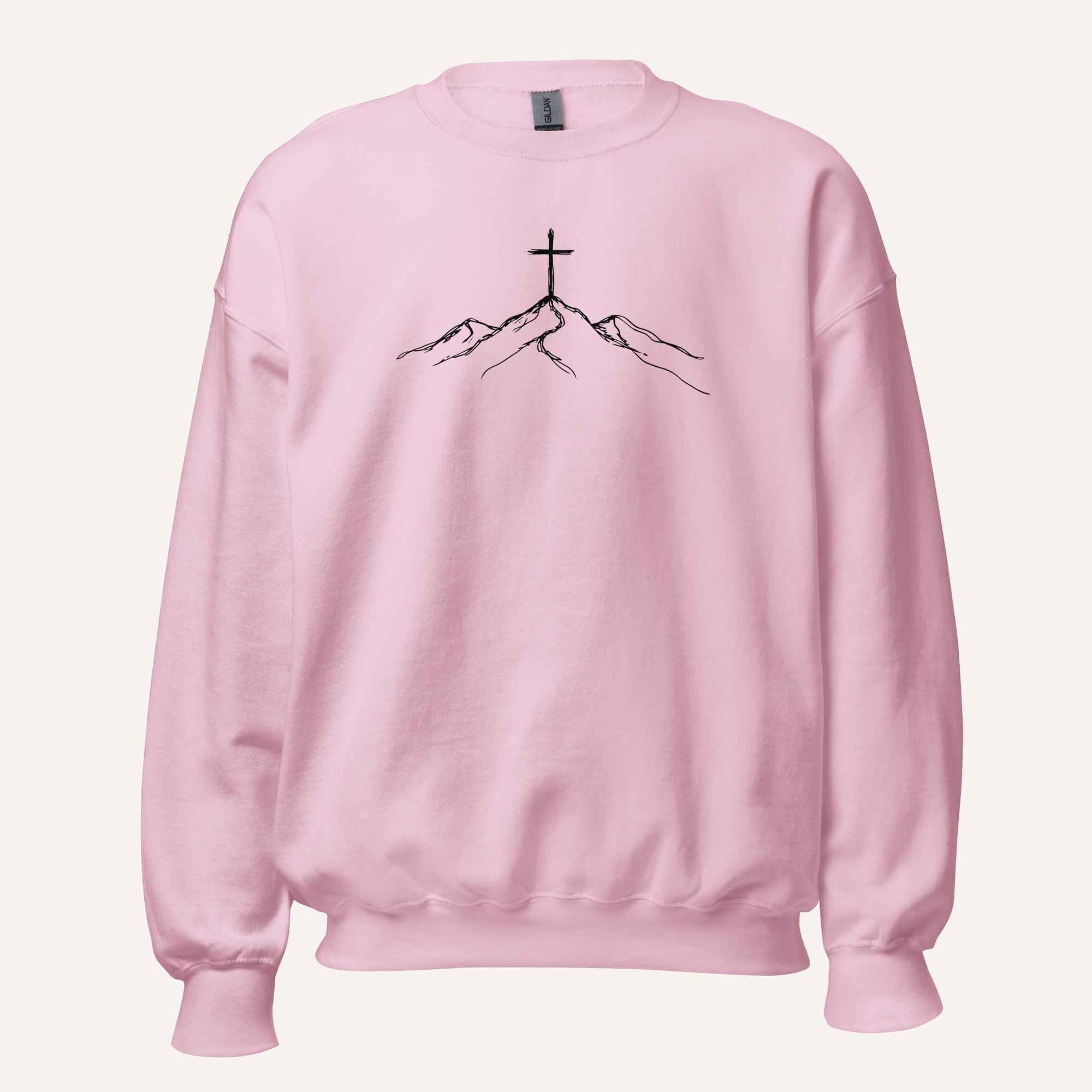 A light pink crewneck sweatshirt featuring a black line art graphic of a cross on top of simple mountains on the chest, displayed against a solid off-white background.