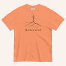 An orange short-sleeve crewneck t-shirt featuring a black line art graphic of a cross on a hill with the word 'Redeemed' printed underneath, displayed on a light cream background.