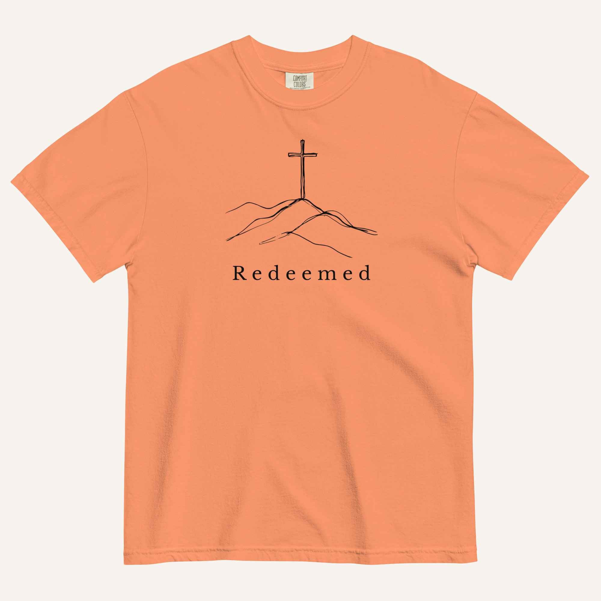 An orange short-sleeve crewneck t-shirt featuring a black line art graphic of a cross on a hill with the word 'Redeemed' printed underneath, displayed on a light cream background.