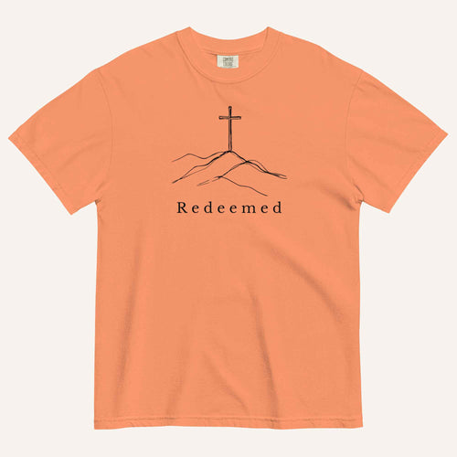 An orange short-sleeve crewneck t-shirt featuring a black line art graphic of a cross on a hill with the word 'Redeemed' printed underneath, displayed on a light cream background.