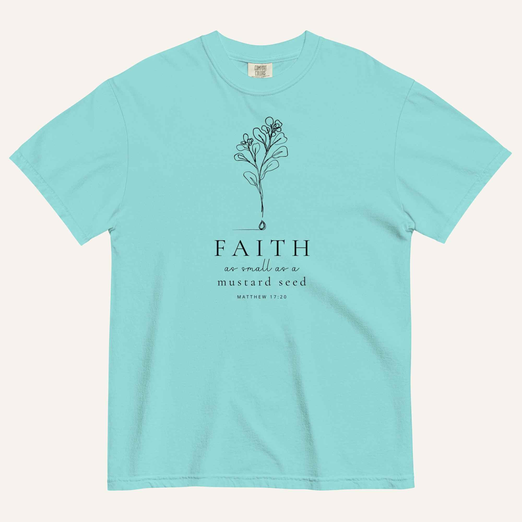 A light blue short-sleeve crewneck t-shirt with a black line-art mustard seed graphic and the text 'FAITH as small as a mustard seed' and 'MATTHEW 17:20' printed on the front, displayed against a light cream background.