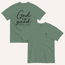 A dark green short-sleeve t-shirt shown from both the front and back on a light cream background. The back features the phrase 'God is good all the time' in black script, and the front has a small black cross on the left chest.