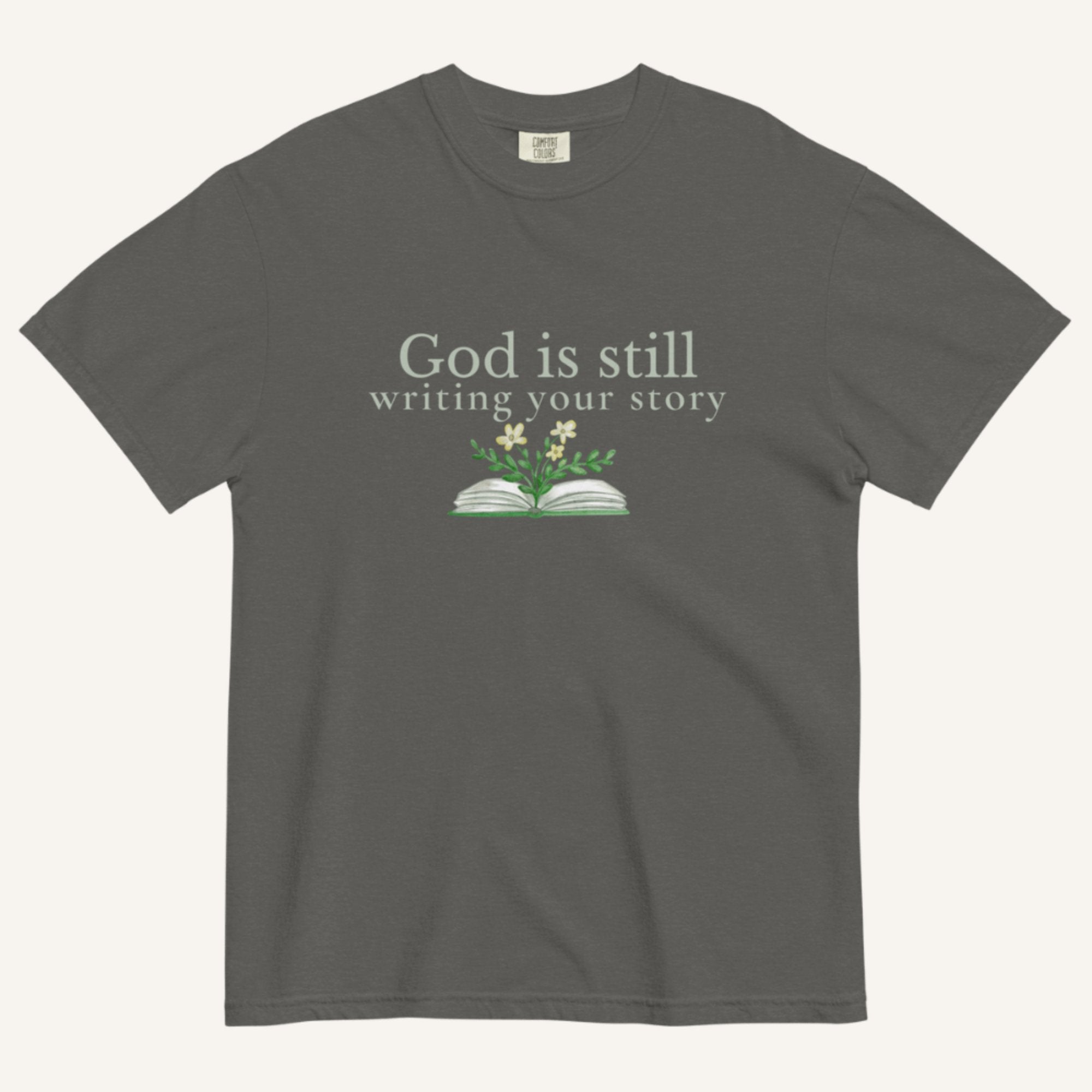 Dark gray t-shirt with 'God is still writing your story' text and book graphic on a white background