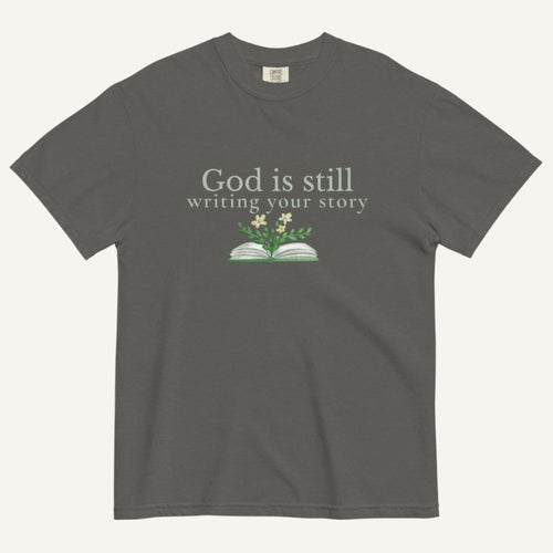 Dark gray t-shirt with 'God is still writing your story' text and book graphic on a white background