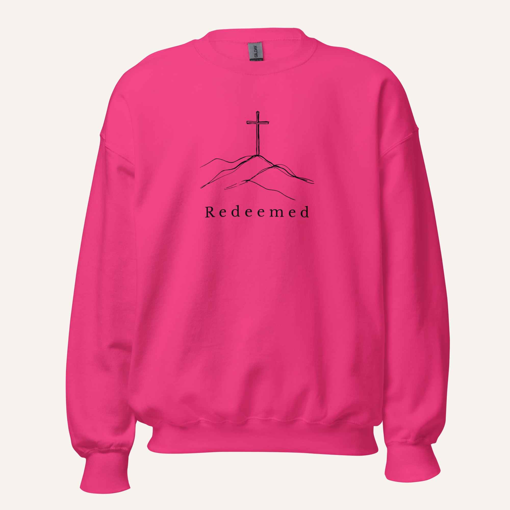A bright pink crewneck sweatshirt featuring a black graphic of a cross on a hill with the word 'Redeemed' printed below it, displayed on a light off-white background.