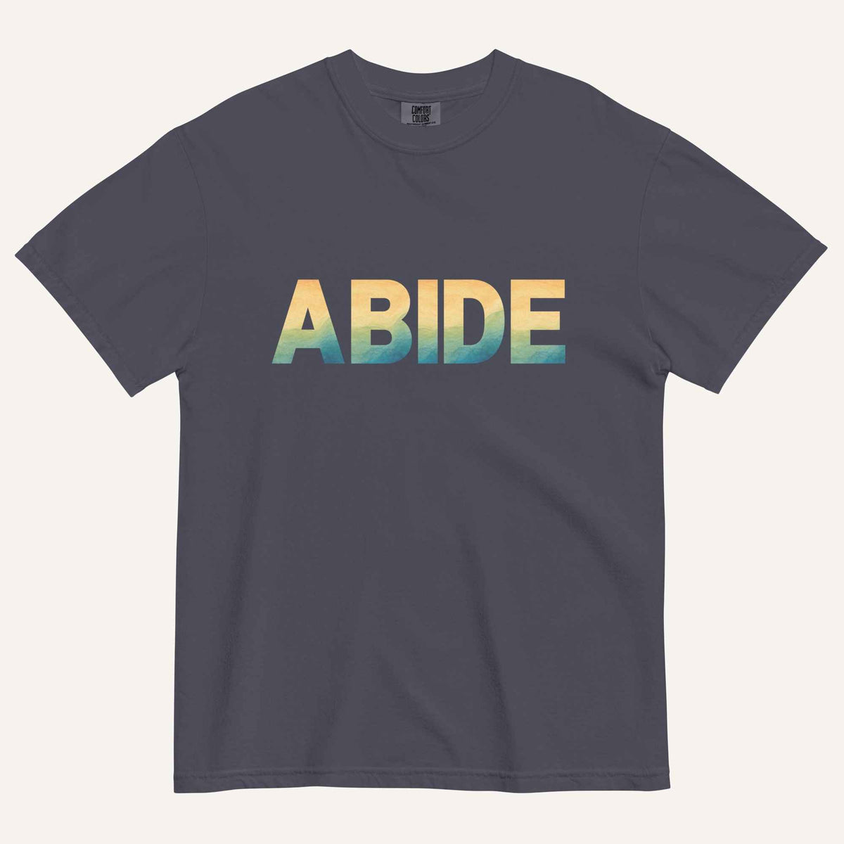 A dark gray crewneck t-shirt featuring the word 'ABIDE' printed across the chest in a bold sans-serif font with a vertical gradient transitioning from yellow to green and blue. The t-shirt is displayed flat against a light cream background.