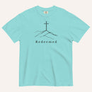 An aqua short-sleeve crewneck t-shirt featuring a black line art graphic of a cross on a hill with the word 'Redeemed' printed underneath, displayed on a light cream background.