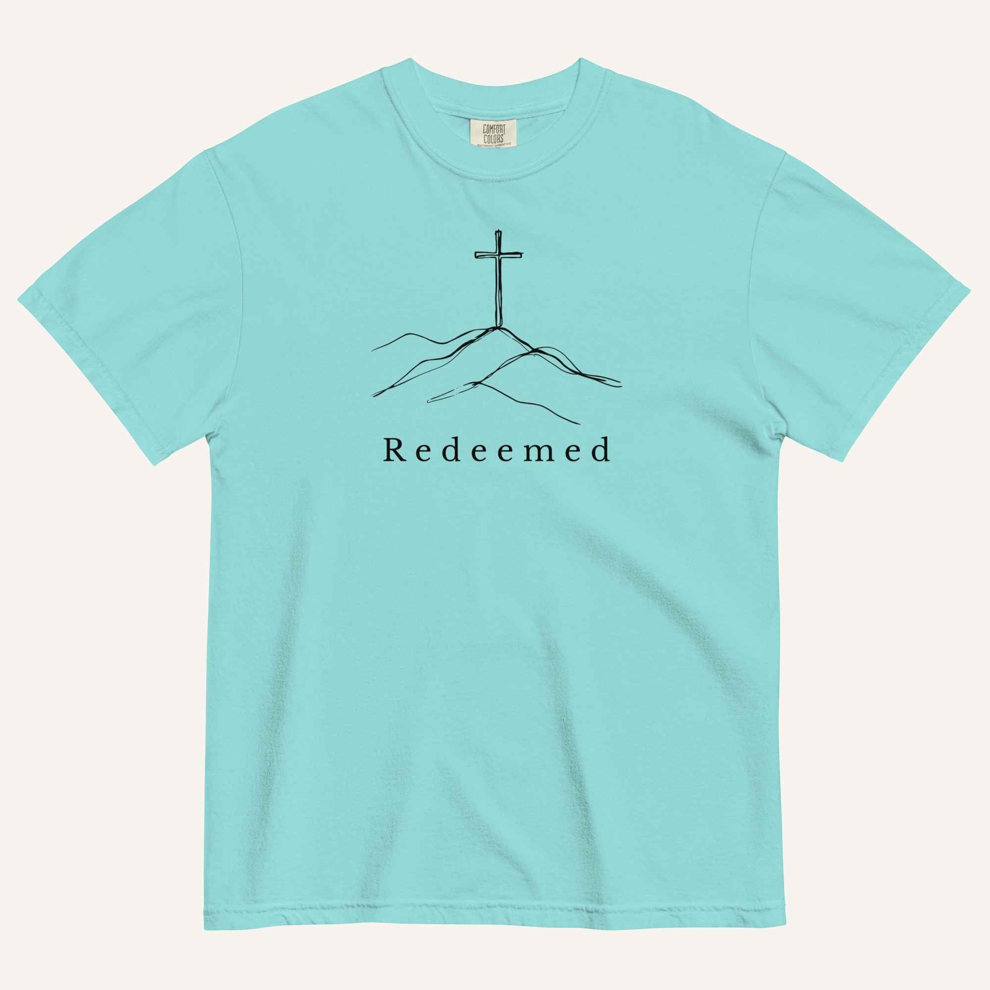 An aqua short-sleeve crewneck t-shirt featuring a black line art graphic of a cross on a hill with the word 'Redeemed' printed underneath, displayed on a light cream background.