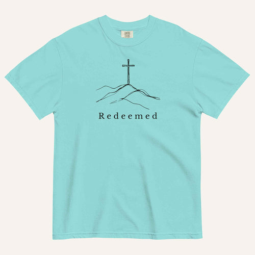 An aqua short-sleeve crewneck t-shirt featuring a black line art graphic of a cross on a hill with the word 'Redeemed' printed underneath, displayed on a light cream background.