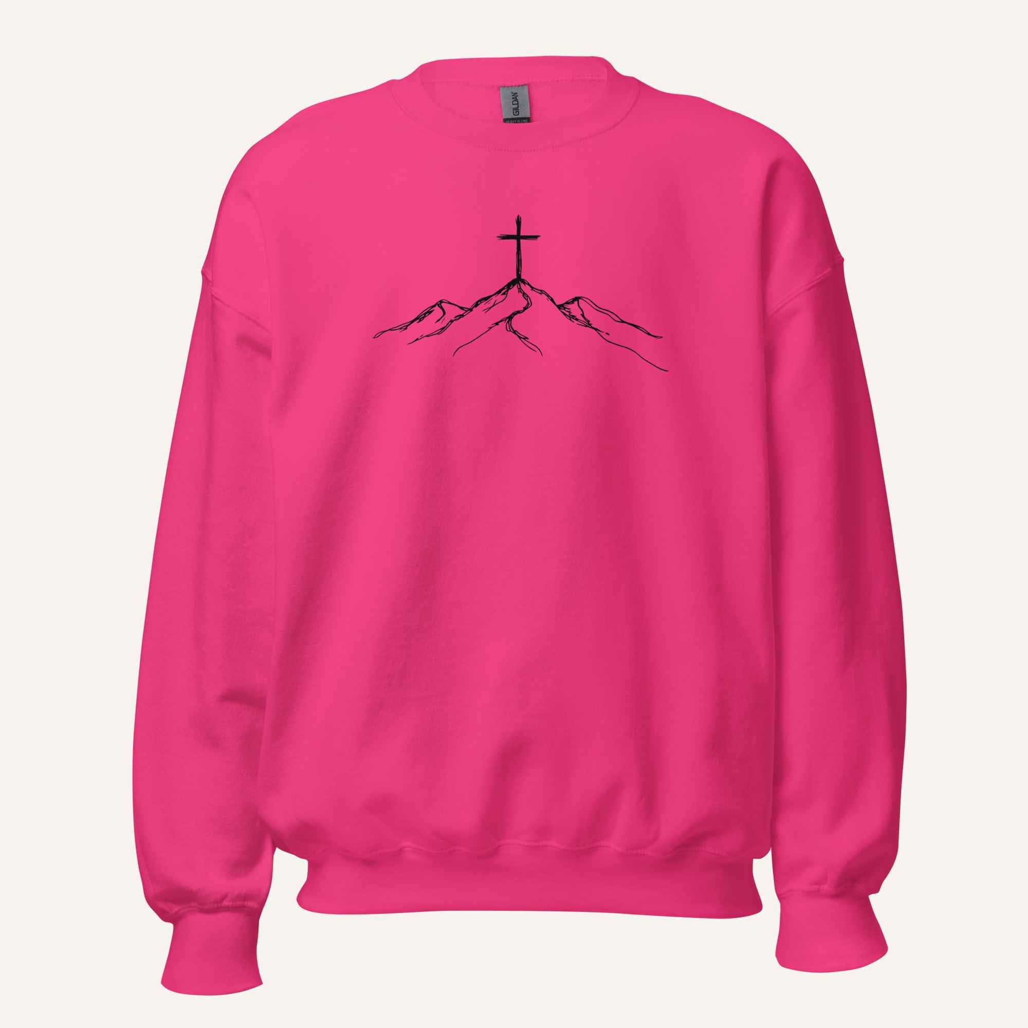A bright pink crewneck sweatshirt featuring a black line art graphic of a cross on top of simple mountains on the chest, displayed against a solid off-white background.