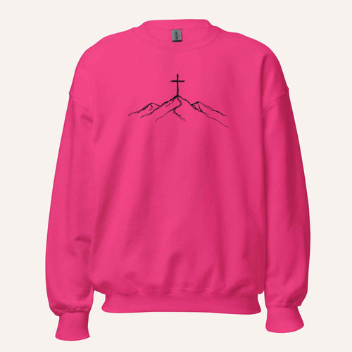A bright pink crewneck sweatshirt featuring a black line art graphic of a cross on top of simple mountains on the chest, displayed against a solid off-white background.
