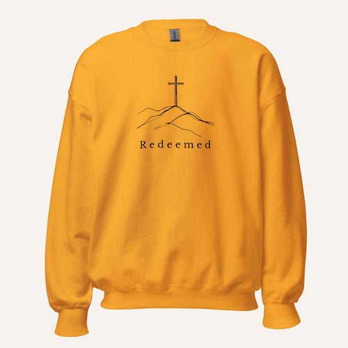 A yellow orange crewneck sweatshirt featuring a black graphic of a cross on a hill with the word 'Redeemed' printed below it, displayed on a clean off-white background.