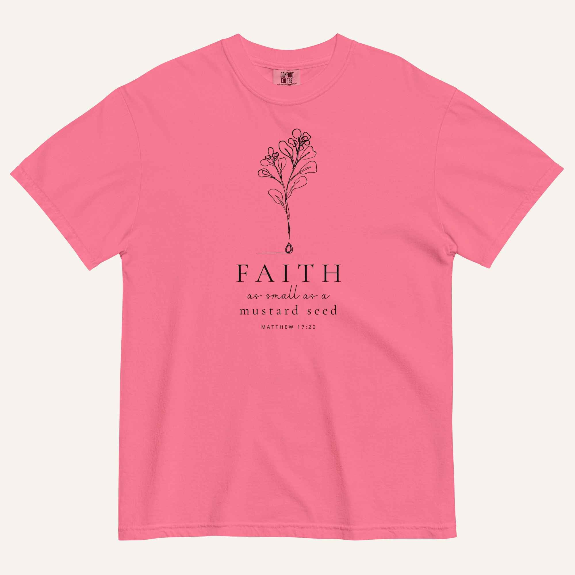A bright pink short-sleeve crewneck t-shirt with a black line-art mustard seed graphic and the text 'FAITH as small as a mustard seed' and 'MATTHEW 17:20' printed on the front, displayed against a light cream background.
