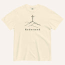 A cream short-sleeve crewneck t-shirt featuring a black line art graphic of a cross on a hill with the word 'Redeemed' printed underneath, displayed on a light cream background.