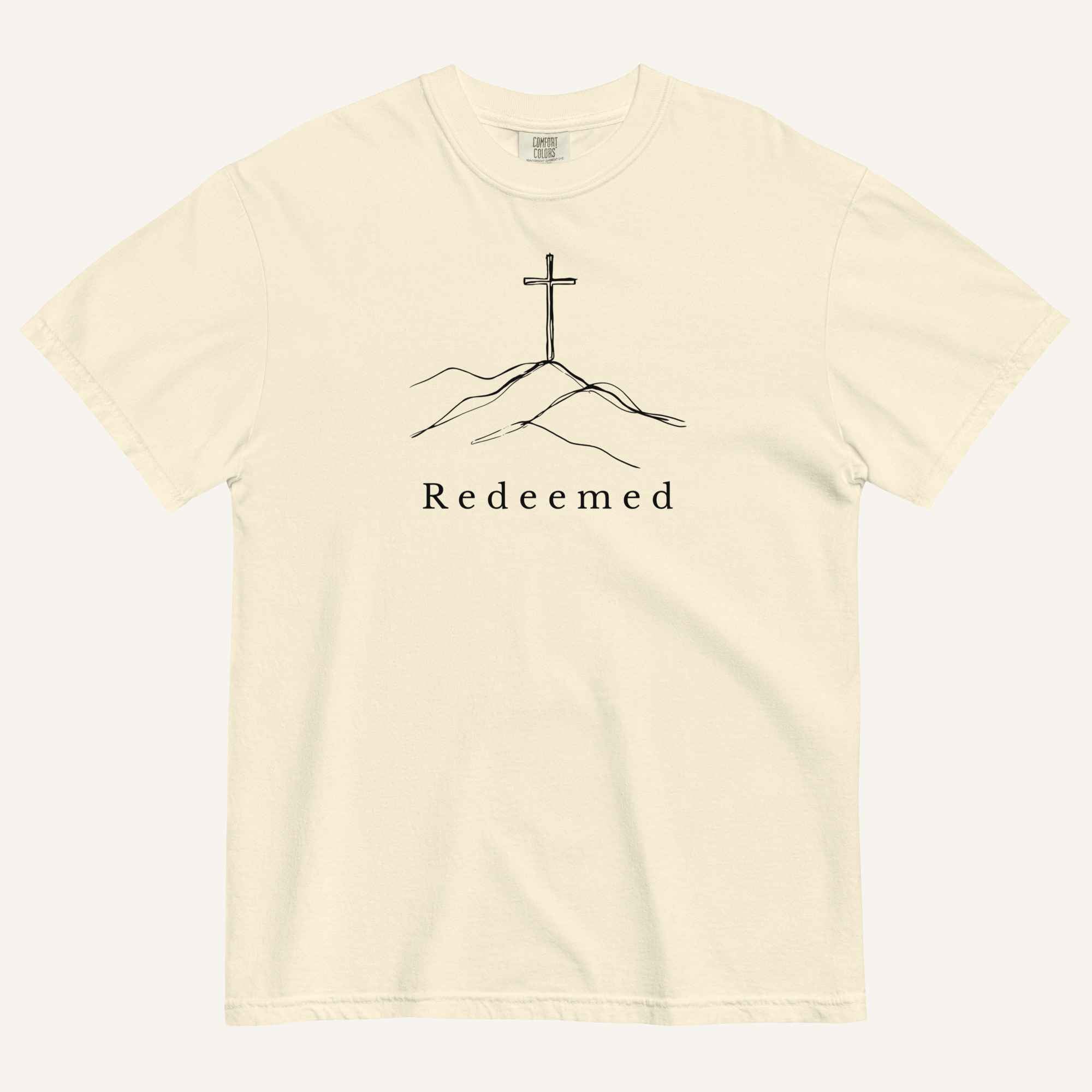 A cream short-sleeve crewneck t-shirt featuring a black line art graphic of a cross on a hill with the word 'Redeemed' printed underneath, displayed on a light cream background.
