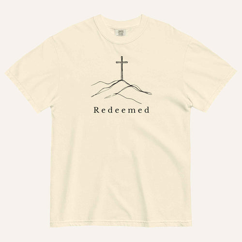 A cream short-sleeve crewneck t-shirt featuring a black line art graphic of a cross on a hill with the word 'Redeemed' printed underneath, displayed on a light cream background.