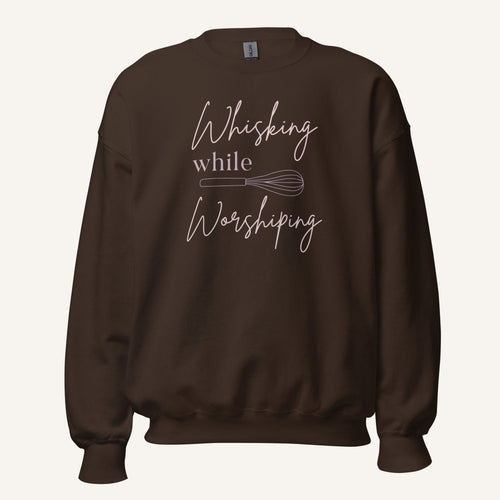 Brown crewneck sweatshirt with "Whisking while Worshiping" pink text with a pink whisk graphic.