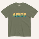An army green crewneck t-shirt featuring the word 'ABIDE' printed across the chest in a bold sans-serif font with a yellow-to-blue gradient, displayed on a light cream background.
