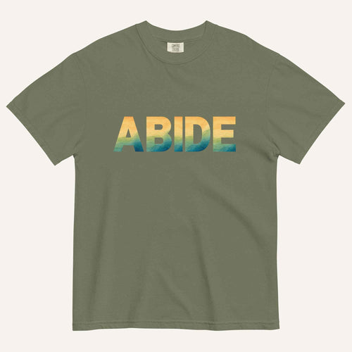 An army green crewneck t-shirt featuring the word 'ABIDE' printed across the chest in a bold sans-serif font with a yellow-to-blue gradient, displayed on a light cream background.