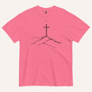 A pink short-sleeve crewneck t-shirt featuring a black line art graphic of a cross on a hill, displayed flat against a light cream background.